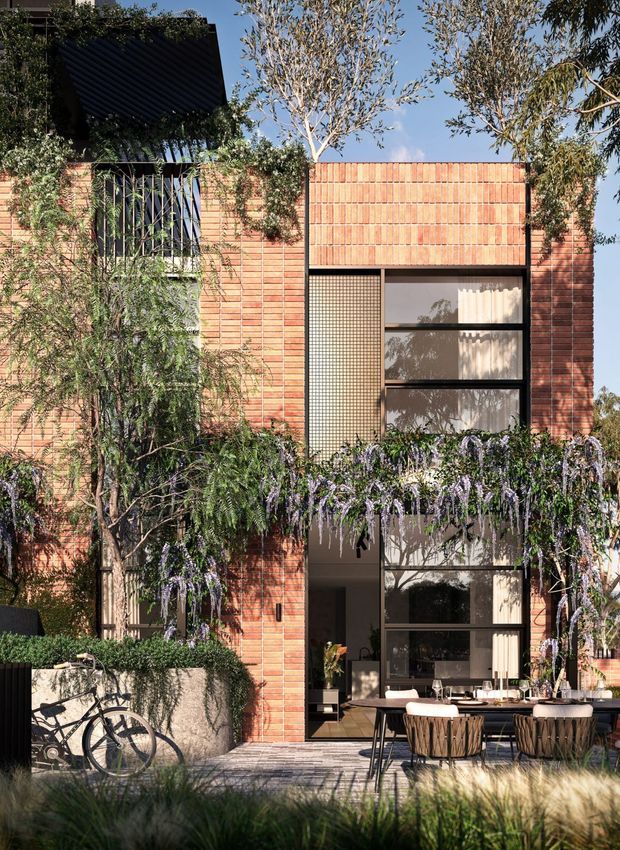 A contemporary multi-storey brick-clad residential facade viewed from a landscaped outdoor area. The building features warm terracotta brickwork, tall black-framed glazing, a projecting dark roof or awning, and a ground-level dining terrace with outdoor furniture. Mature planting, climbing greenery, and a bicycle soften the architecture.
