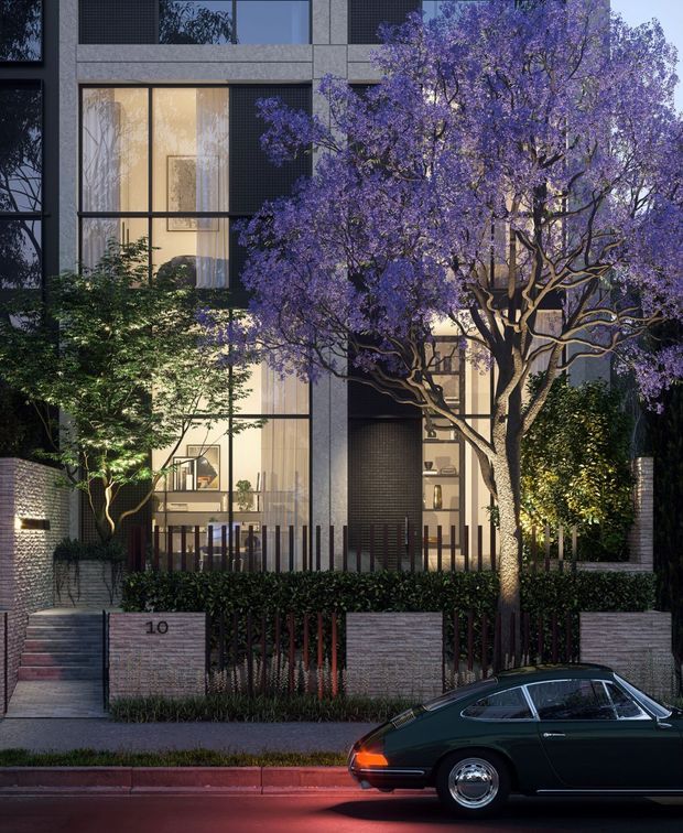 Street-level dusk view of a contemporary multi-storey residential building with a dark grey masonry or concrete facade, large floor-to-ceiling windows, warm interior lighting, and a landscaped frontage. A prominent purple-flowering tree partially obscures the facade, while low brick retaining walls, hedges, fencing, and entry steps define the property boundary. A parked vintage car is visible in the foreground.