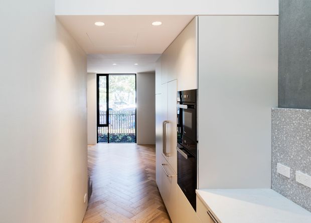 A narrow contemporary kitchen corridor with white full-height cabinetry, integrated black appliances, a pale stone benchtop, and a grey speckled splashback. Warm timber parquet flooring leads toward a glazed rear door, while recessed ceiling lights and minimal detailing create a clean, streamlined interior.