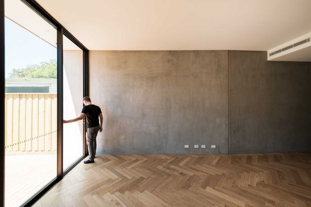 A mostly empty contemporary interior with a full-height black-framed glazed sliding door opening to an exterior deck or balcony. The room features a large smooth grey concrete-look feature wall, warm herringbone timber flooring, a simple white ceiling with linear air-conditioning ventilation, and minimal electrical outlets. A person stands by the glazing, emphasizing the generous opening and natural light.
