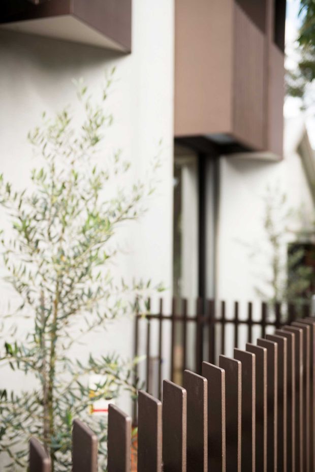 Close-up exterior view of a contemporary residential facade, featuring white rendered walls, projecting dark brown upper volumes, narrow vertical glazing, and a dark timber-toned vertical slat fence in the foreground. Soft landscaping partially screens the facade.