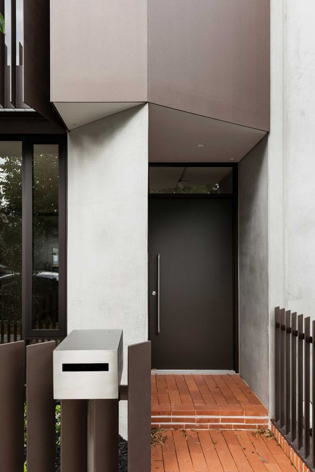Close-up view of a contemporary residential entry facade. A dark charcoal front door with a long vertical pull handle is recessed beneath a projecting angular canopy, framed by pale rendered or concrete surfaces and dark metal glazing. Red brick steps and paving lead to the entrance, with a minimalist metal letterbox and vertical metal balustrade or fence in the foreground.
