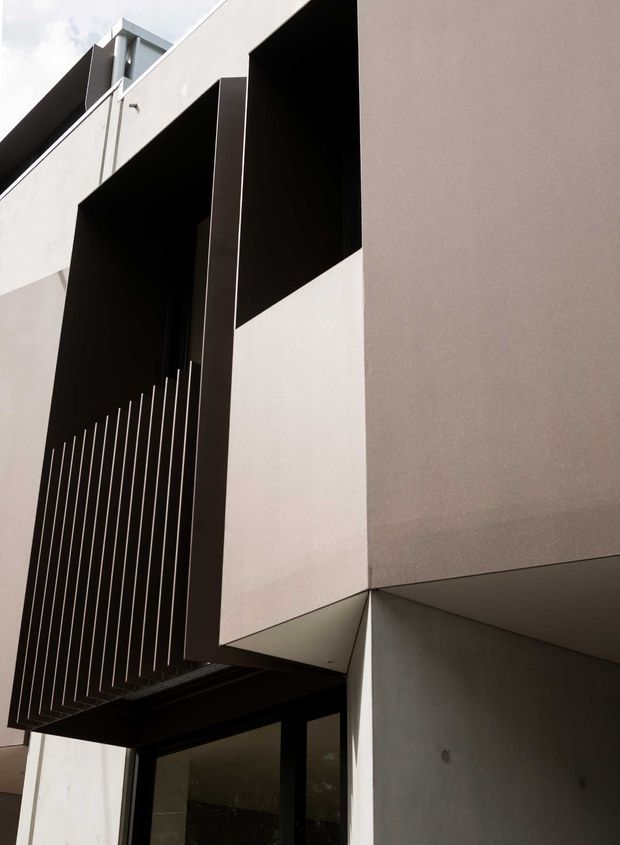Close-up upward view of a modern multi-level building facade with strong geometric forms, projecting pale rendered volumes, deep dark recessed openings, and a tall vertical metal slat screen over glazing. The composition is minimalist and angular, with contrasting light and charcoal surfaces and a sheltered entry or lower-level opening.
