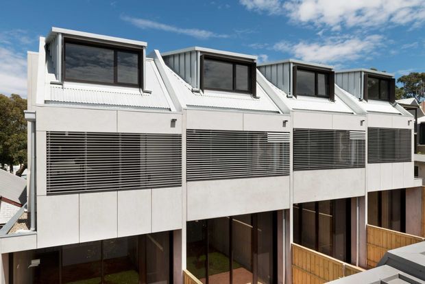 Exterior view of a contemporary multi-unit residential building with a repeated terraced form. The pale facade features projecting upper dormer-like volumes, dark-framed windows, extensive horizontal external sun-shading louvres, and private timber-fenced ground-level courtyards. The building appears multi-storey and densely arranged in an urban or suburban setting beneath a blue sky.