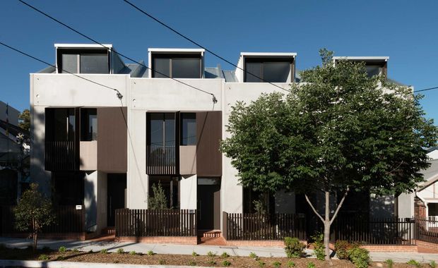 Street-facing view of a contemporary multi-storey residential townhouse development with a flat-roofed, rectilinear facade. The building uses pale rendered/concrete-like wall surfaces, dark window and door frames, projecting box-like upper elements, balconies or screened openings, and dark vertical metal balustrades. A mature street tree partially obscures the right side, with front fences, paving, and neighbouring homes visible.