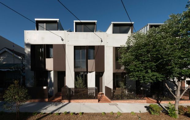 Street-facing exterior view of a row of attached contemporary townhouses. The facade combines pale rendered concrete-like walls, dark vertical window and screen elements, projecting upper-level box forms, and rooftop clerestory or dormer-like structures. Small fenced front courtyards, brick-edged paths, street trees, and overhead utility wires are visible.