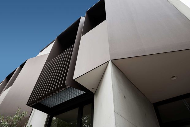 Upward-looking architectural exterior view of a contemporary multi-level building. The facade combines smooth pale rendered or concrete planes with sharply projecting geometric volumes, deep recessed openings, dark vertical fins, and a dark glazed entrance canopy. A clear blue sky and minimal landscaping are visible at the edges.