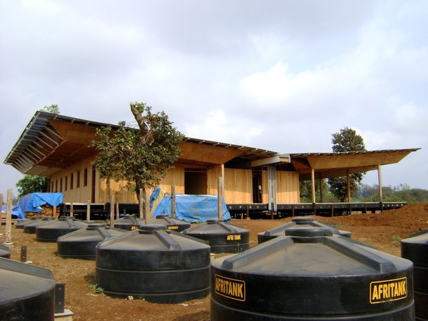 A low-rise timber-clad building under construction, featuring broad asymmetrical roof overhangs, exposed timber framing, and elevated sections. Large black water storage tanks occupy the foreground, with construction materials and earthworks surrounding the structure.