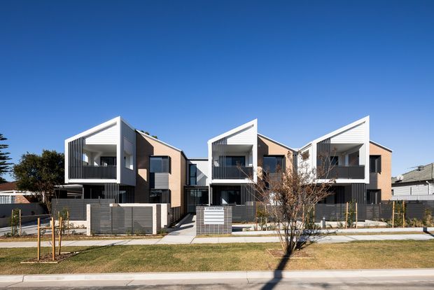 Street-facing view of a contemporary multi-unit residential development with three attached townhouses. The buildings feature stepped geometric forms, projecting upper-level box volumes, pale rendered walls, darker vertical cladding, recessed balconies, and enclosed front courtyards with horizontal metal fencing. A landscaped verge and sidewalk sit in the foreground under a clear blue sky.