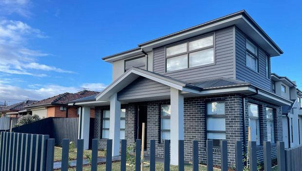 Two-storey contemporary suburban house viewed from the street, featuring dark grey brickwork on the lower level, grey horizontal weatherboard cladding above, a low-pitched roof with broad eaves, large windows, and a covered front entry. A dark vertical-slat fence frames the foreground.