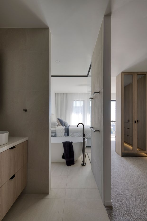 A contemporary bathroom viewed from a hallway or dressing area, with large pale stone-look wall and floor tiles, a timber vanity, freestanding white bathtub, and matte black tapware. The bathroom opens directly onto a bedroom visible in the background, which has a neatly made bed and sheer curtains. To the right is a built-in timber wardrobe with mirrored doors and subtle under-cabinet lighting. The space has a restrained, warm-neutral palette and clean-lined minimalist detailing.