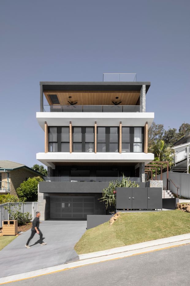 Street-facing view of a contemporary multi-storey residence on a sloped suburban site. The home has a flat roof, broad projecting balconies, dark framed glazing, white rendered horizontal bands, exposed brick piers, a timber-lined upper balcony ceiling, stone-clad vertical elements, and an integrated garage. Landscaped terraces and neighbouring homes surround the property.