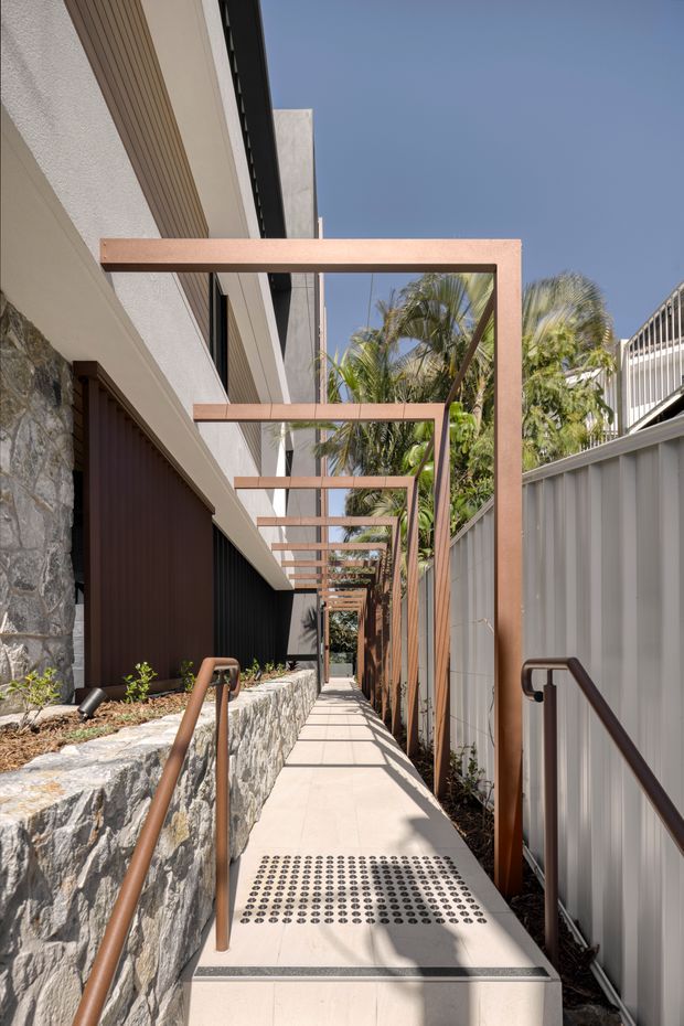 A narrow landscaped external walkway runs alongside a contemporary building, with a pale paved path, accessibility handrails, tactile indicators, low stone retaining wall, and repeated rust-coloured rectangular overhead frames. Textured white walls, dark cladding, corrugated metal fencing, tropical palms, and bright sunlight create a modern residential or commercial approach.