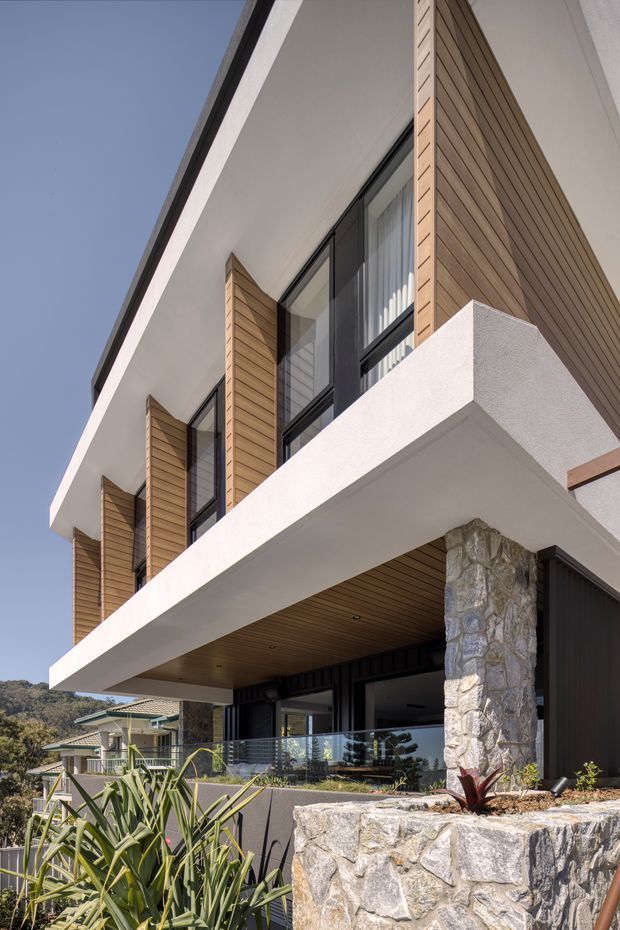 Upward-looking exterior photograph of a contemporary multi-level residence with a strong projecting white concrete balcony and roof overhang. The facade combines warm horizontal timber cladding, dark-framed glazing, white rendered surfaces, and substantial rough natural-stone columns and retaining walls. A glass balustrade, recessed entry or outdoor living area, and landscaped succulent planting are visible, with neighbouring houses and a wooded hillside in the background.