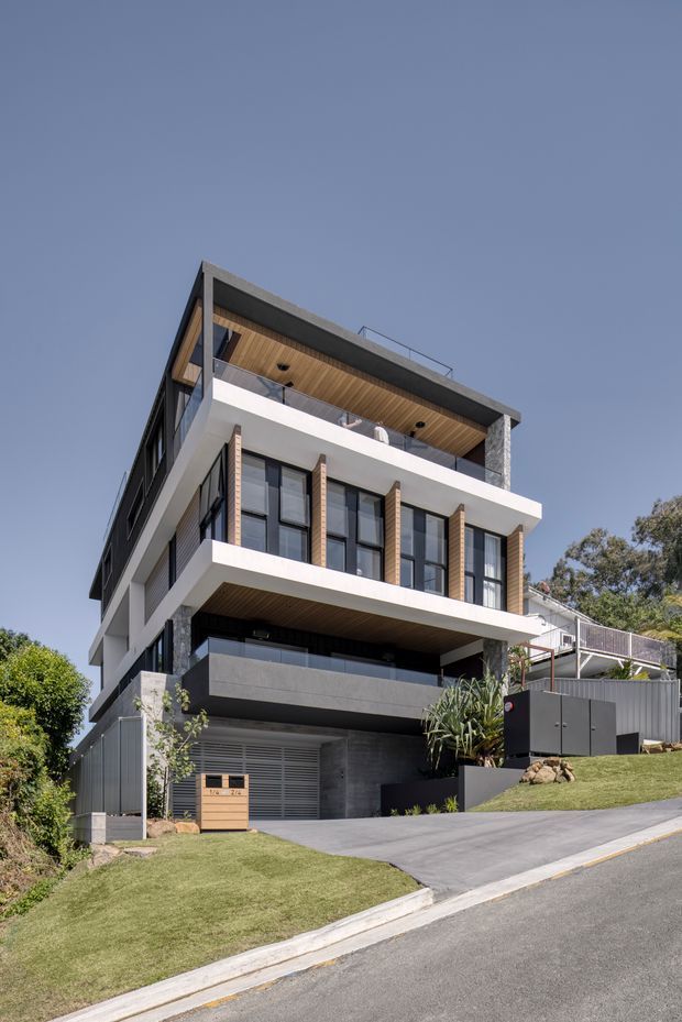 Street-level exterior photograph of a contemporary multi-storey residence on a sloping suburban site. The building has a strongly geometric facade with white projecting slabs, dark grey wall surfaces, extensive black-framed glazing, vertical timber-look screens, a timber-lined upper balcony ceiling, a flat roof, and an integrated street-facing garage. A landscaped lawn and driveway sit in front of the house.