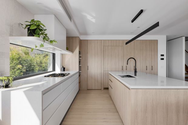 Bright contemporary kitchen with light timber cabinetry, white stone benchtops, integrated appliances, a large central island with sink, and expansive side glazing overlooking greenery. Minimal black linear pendant lights and a black tap provide contrast against the pale neutral finishes.