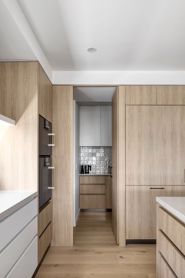 A compact contemporary kitchen with a narrow walk-in pantry or scullery opening at the centre. The room features extensive light timber-grain cabinetry, integrated dark appliances, pale stone benchtops, warm timber flooring, and a reflective square-tile splashback in the pantry. White walls and ceiling with recessed lighting create a clean, streamlined look.