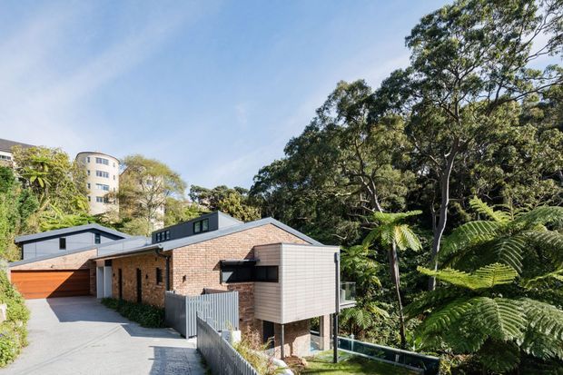 Exterior view of a contemporary multi-level brick house set into a lush, heavily vegetated urban or suburban site. The building has a pitched gable roof with dark metal roofing, projecting box-like additions clad in horizontal timber-toned boards, large glazed openings, and an integrated garage accessed by a concrete driveway. Dense mature trees and tree ferns surround the property, with a taller apartment or residential building visible in the background.