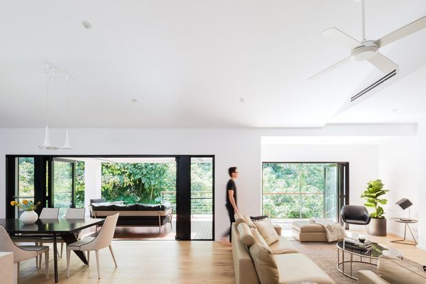 Bright open-plan residential interior combining a dining area and living room. The space features white walls and ceiling, pale timber flooring, expansive black-framed glazed sliding doors opening to greenery, a large sectional sofa, neutral lounge furniture, a contemporary dining table with chairs, recessed lighting, and a ceiling fan. The room has a clean, airy, contemporary feel.
