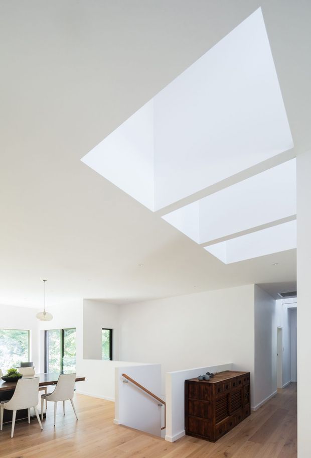 Bright contemporary open-plan interior with a dining area, staircase, hallway, and timber floors. Large rectangular skylights cut through the white ceiling, creating a dramatic overhead feature and abundant natural light. The space includes white walls, slim dark-framed windows, a white dining table and chairs, a timber stair handrail, and a dark timber sideboard.