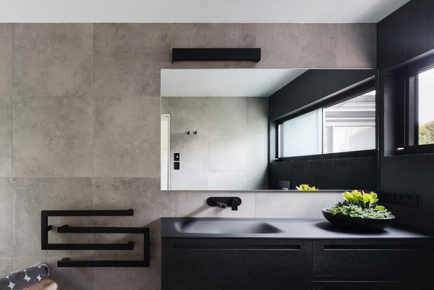 Contemporary bathroom vanity with a large frameless mirror, dark wall-mounted double basin and cabinetry, black wall-mounted tapware, concrete-look wall tiles, and a sculptural black towel rail. A horizontal window brings in natural light, while a small planter adds greenery.