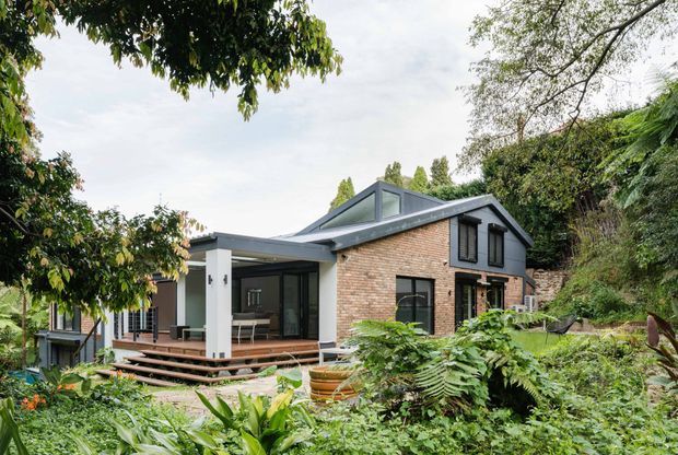A contemporary two-storey brick house set within dense established garden landscaping. The home has a pitched gable roof with dark metal roofing, large glazed openings, and a covered outdoor living area opening onto a timber deck with steps. Lush planting surrounds the house, while a sloped, vegetated site rises behind it.