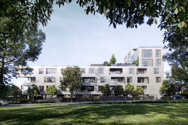 Exterior architectural rendering of a contemporary multi-storey apartment building set behind landscaped lawn and mature trees. The light-coloured facade has varied volumes, recessed balconies, large rectangular windows, and dark balcony railings, with a taller section rising at the right.