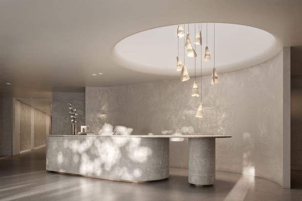 A refined, minimalist reception or kitchen-like counter space with a long curved stone island, cylindrical support, textured pale walls, and a dramatic oval recessed ceiling opening. Multiple slender pendant lights hang over the counter, while soft daylight creates dappled shadows across the surfaces. The palette is warm neutral grey and beige with a calm, luxurious atmosphere.