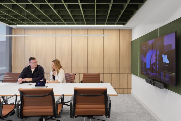 A contemporary commercial meeting room with a long white conference table, brown leather office chairs, extensive light timber built-in wall cabinetry, and a large wall-mounted video display. The room features a dark exposed grid ceiling, grey carpet, and a green acoustic feature wall around the screen.
