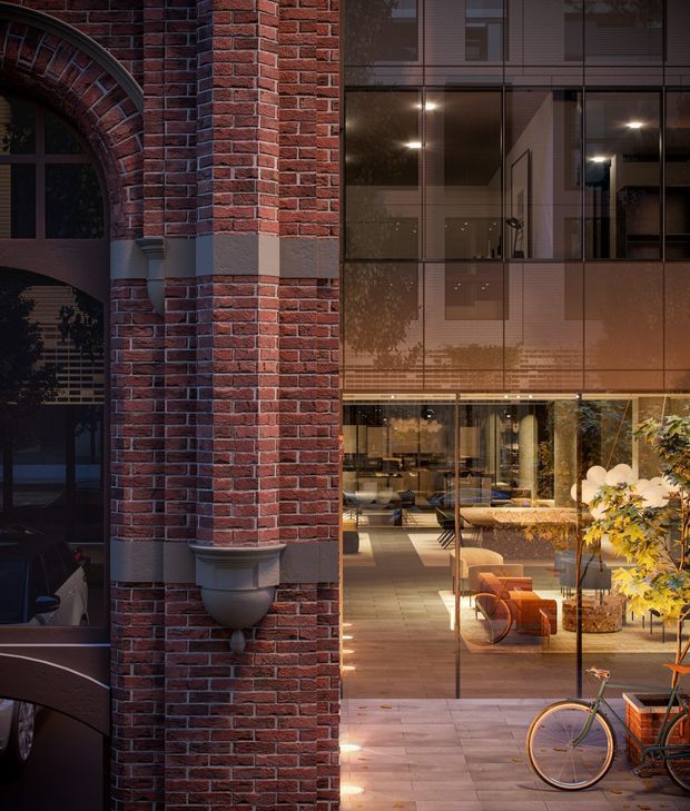 A dusk architectural view juxtaposing a heritage-style red brick facade with arched openings and decorative masonry details against a contemporary glazed addition. Through the floor-to-ceiling glazing, a warmly lit interior with dining and lounge furniture, pendant lighting, tiled flooring, and indoor planting is visible. Bicycles appear in the foreground, while reflections overlay parts of the glass facade.