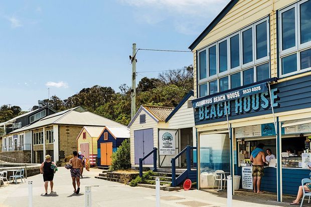 Sunny exterior streetscape of a coastal beach-house-style commercial building, with blue-and-white weatherboard cladding, large upper-level windows, a prominent “Beach House” sign, adjacent small painted timber buildings, pedestrian paving, and outdoor seating.