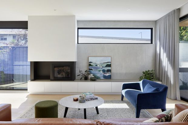 Bright contemporary living room with a minimalist built-in fireplace wall, pale textured plaster finish, low white cabinetry, expansive glazing, full-height curtains, and a deep blue sculptural armchair. A neutral rug, round coffee table, green ottoman, artwork, and indoor plants add restrained colour and warmth.