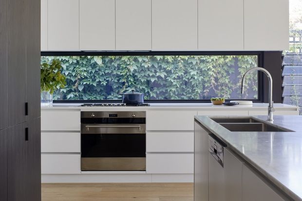 Contemporary kitchen interior with white handleless cabinetry, integrated wall storage, a built-in stainless-steel oven, and a large island with a grey stone benchtop and double sink. A full-width window above the rear bench overlooks dense greenery, while timber flooring and a tall curved chrome tap add warmth and contrast.