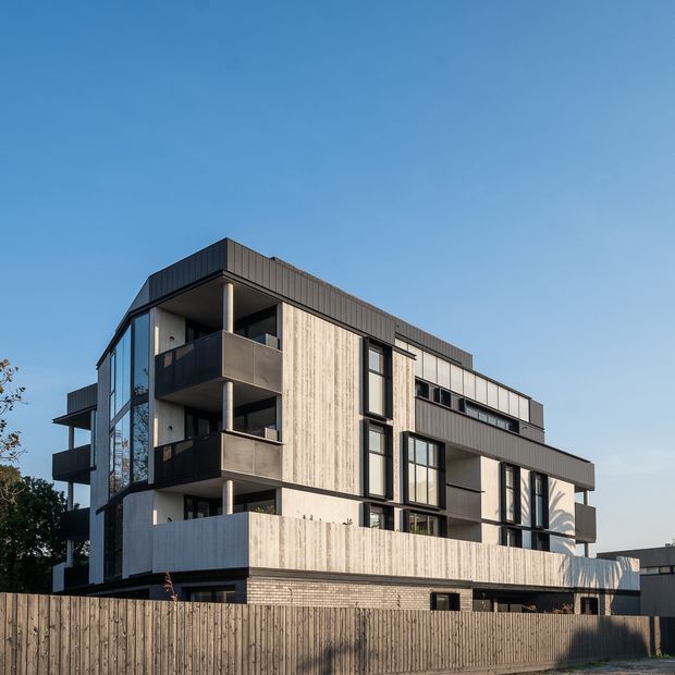 Exterior view of a contemporary multi-storey residential apartment or townhouse building with a boxy flat-roofed form. The facade combines dark vertical cladding, pale textured concrete or fibre-cement panels, extensive black-framed glazing, projecting balconies, and a timber privacy fence in the foreground. The building is photographed from the street against a clear sky.