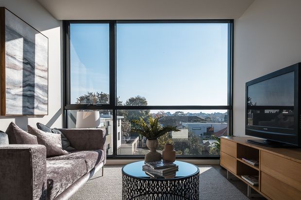 A bright contemporary living room with a large black-framed picture window overlooking a suburban landscape. The room contains a textured sectional sofa, round glass-top coffee table with an open metal base, wall art, potted plants, and a timber media console with a large television. Neutral finishes and abundant daylight create an airy, refined atmosphere.