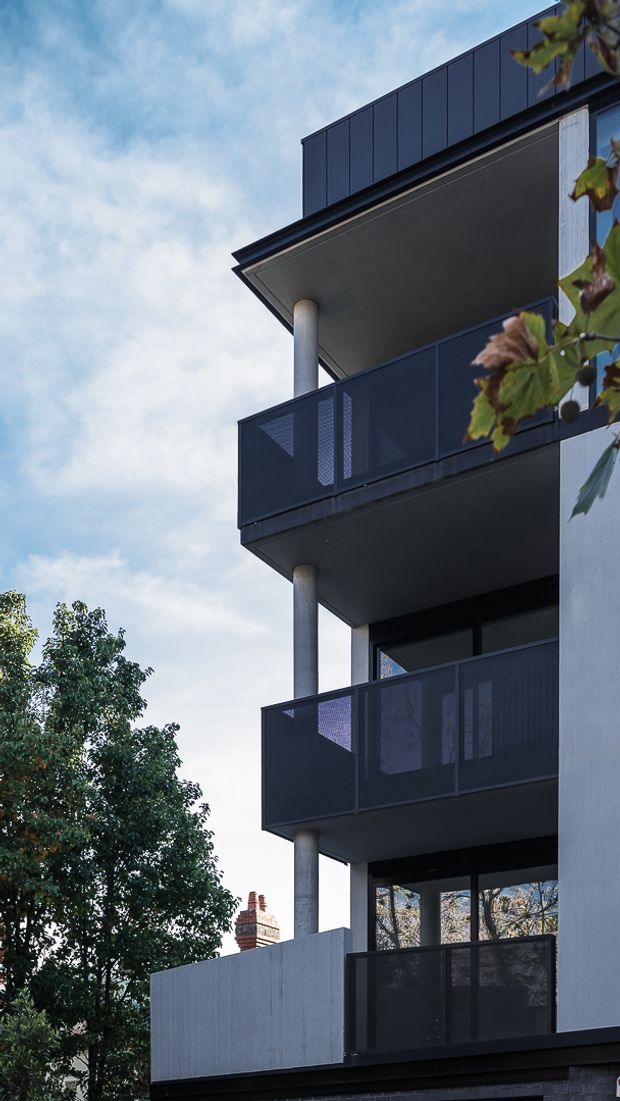 Vertical exterior architectural photograph of a contemporary multi-storey residential building, viewed upward along a stack of projecting balconies. The facade combines dark metal-framed screened balustrades, pale rendered walls, exposed round concrete columns, deep balcony soffits, and large dark-framed glazing. Mature trees and a neighbouring brick chimney are visible around the building, beneath a bright partly cloudy sky.