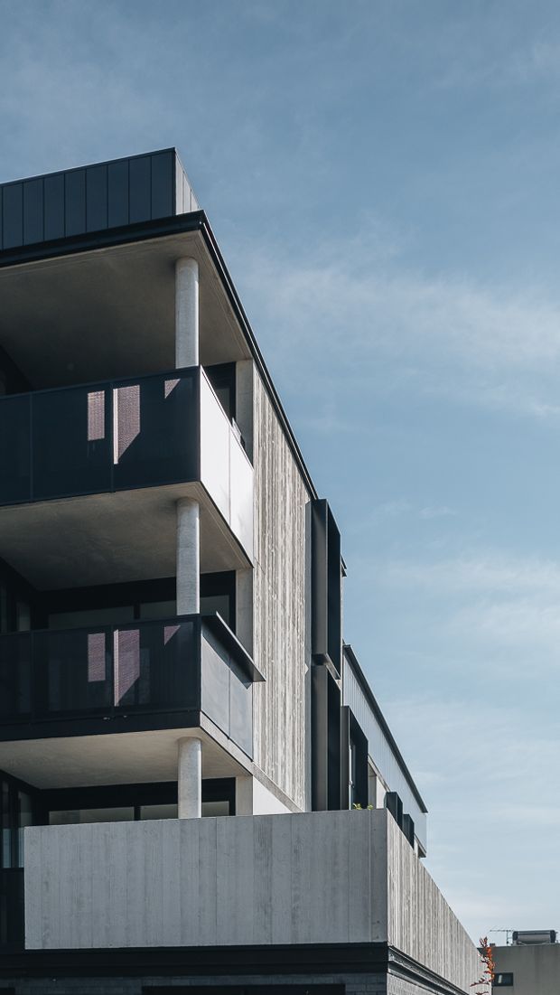 Vertical architectural photograph of a contemporary multi-storey apartment building viewed from below. The facade combines dark metal balcony balustrades and roof elements with exposed concrete slab balconies, cylindrical concrete columns, and a tall vertically textured concrete or cement-clad wall. Repeated balconies and strong geometric lines dominate the composition against a pale blue sky.