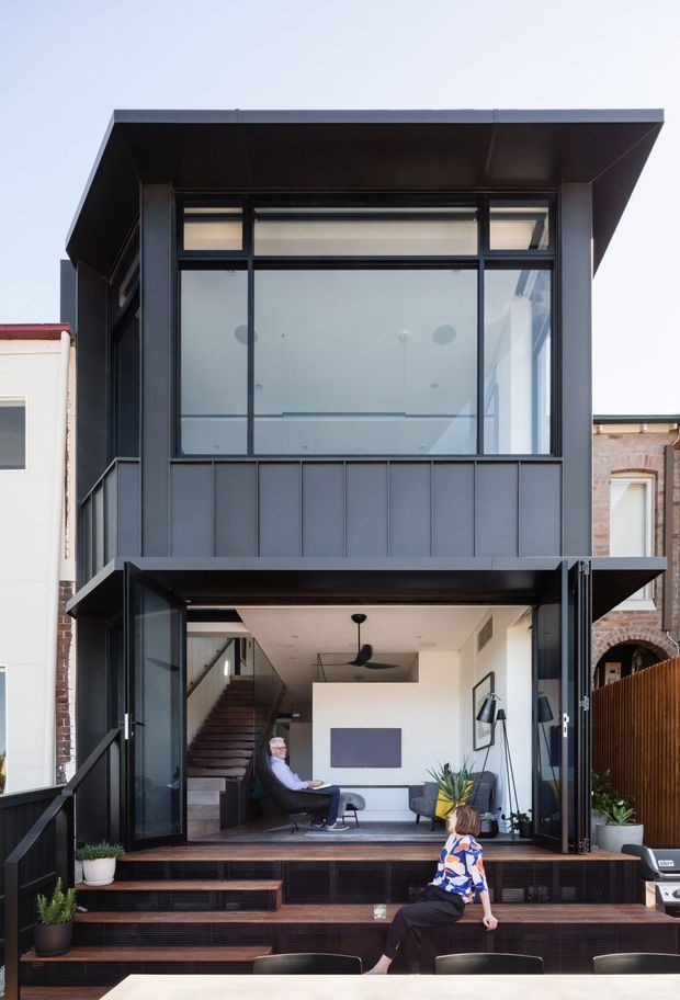 A narrow, multi-storey urban residence with a contemporary black metal facade, large glazed openings, and a projecting flat roof. The open ground-level living area connects to a stepped timber deck, while an internal staircase and compact seating area are visible inside. Dark vertical cladding and framing contrast with white interior walls and neighbouring brick buildings.