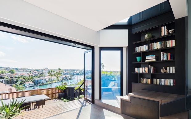 Bright contemporary living room with floor-to-ceiling glazing opening onto a timber deck and expansive waterfront view. A dark built-in wall unit with open bookshelves, integrated cabinetry and a bench seat forms the main interior feature. The room has a pale ceiling and walls, dark window frames, timber flooring, and strong indoor-outdoor connection.