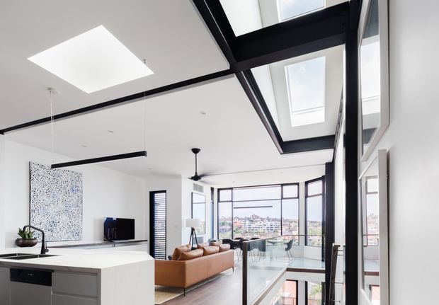 Bright contemporary open-plan interior viewed from a kitchen island, with a white kitchen, timber-toned flooring, adjoining living area, extensive black-framed glazing, skylights, a glass balustrade, and a visible lower-level void or stair opening. The space is flooded with natural light and overlooks a dense urban neighbourhood.