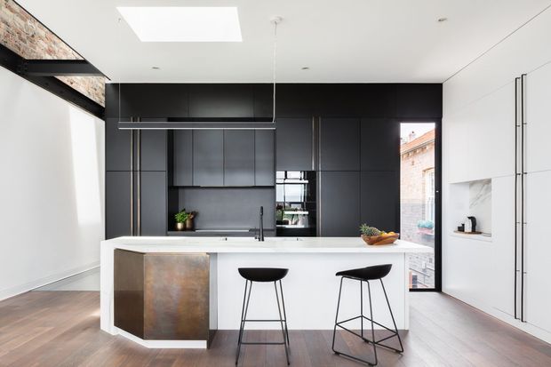 A contemporary open-plan kitchen with a large white island and two black bar stools. The kitchen features full-height matte black cabinetry, integrated appliances, a dark grey benchtop area, a slim linear pendant light, and a skylight. Warm timber flooring contrasts with the monochrome joinery. An exposed brick wall and black steel beam are visible at the left, while a tall window and open shelving bring in daylight.