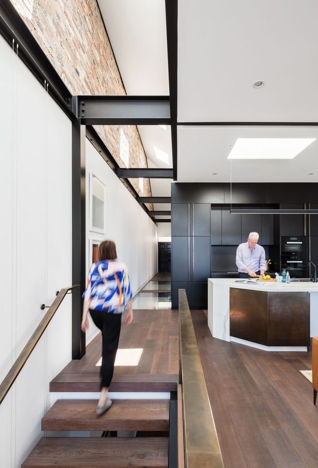 A contemporary open-plan kitchen and circulation space with a compact stairway in the foreground. Dark timber flooring, black steel structural beams and trims, exposed brickwork, white wall panels, and extensive dark built-in kitchen cabinetry create an industrial-modern interior. A white island with a dark metal-clad side occupies the kitchen, while a raised walkway and skylights bring light into the long narrow space.