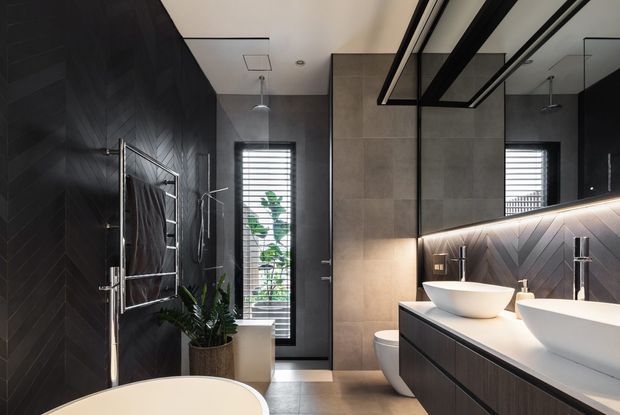 A sophisticated contemporary bathroom featuring a double vanity with white vessel basins, dark timber-look chevron wall panelling, large grey wall tiles, black-framed mirrors, chrome tapware, a heated towel rail, and a freestanding bathtub partially visible in the foreground. Natural light enters through a narrow louvred window, complemented by warm under-mirror lighting.