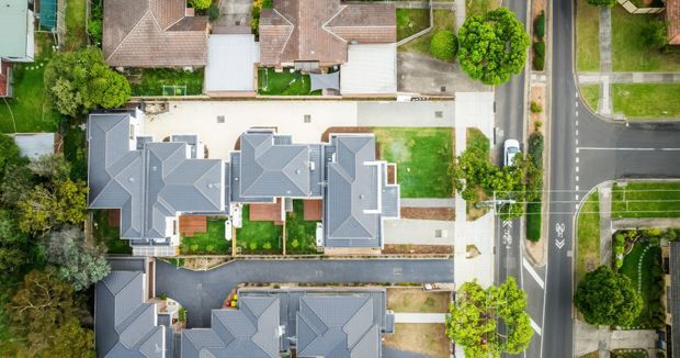 Aerial top-down view of a suburban residential development, showing multiple attached or closely spaced homes with grey pitched roofs, driveways, paved access lanes, small lawns, established trees, and surrounding streets.