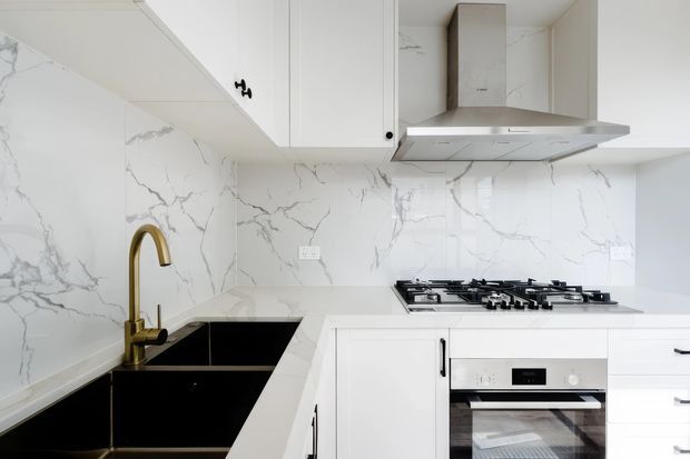 Bright contemporary white kitchen with full-height white marble-look splashback and matching benchtops, white overhead and base cabinetry, a black double-bowl sink, brass gooseneck tap, stainless steel rangehood, gas cooktop and built-in oven.