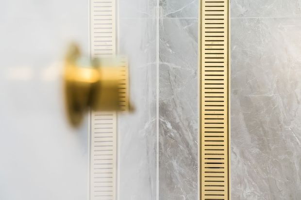 Close-up architectural detail of a luxurious bathroom wall, featuring grey marble-effect stone surfaces, a brushed gold vertical slotted drain or trim, and a blurred gold-toned wall fixture.