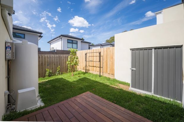 A compact private backyard courtyard with a timber decking platform, lawn, timber boundary fencing, a small young tree, and a freestanding light-coloured outbuilding or garage with dark grey double doors. Modern neighbouring homes are visible beyond the fence under a bright blue sky.