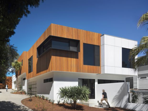 Exterior view of a contemporary multi-storey residence with a strong geometric composition. The upper level is clad in warm vertical timber battens, contrasted with smooth white rendered and panelled walls, dark-framed glazing, and projecting box-like window volumes. A paved pedestrian approach, landscaped garden beds, boundary fencing, and a partially visible neighbouring structure frame the house.