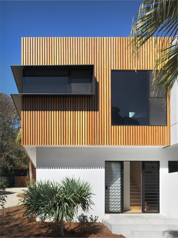 Exterior view of a contemporary multi-storey residence with a white ground floor and upper facade clad in closely spaced vertical timber battens. The facade features large dark-framed windows, a projecting box window, a recessed entry with timber-lined stairwell, and clean geometric forms. Drought-tolerant landscaping and a palm are visible in the foreground under a clear blue sky.