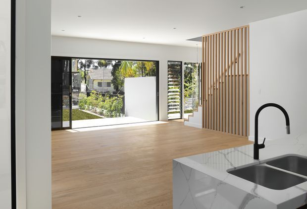 Bright open-plan interior viewed from the kitchen, with a marble-look island and black gooseneck tap in the foreground. The main living area has pale timber flooring, white walls and ceiling, large black-framed glazed doors, and a vertical timber slat screen beside a compact stair. A landscaped garden and neighbouring house are visible through the glazing.